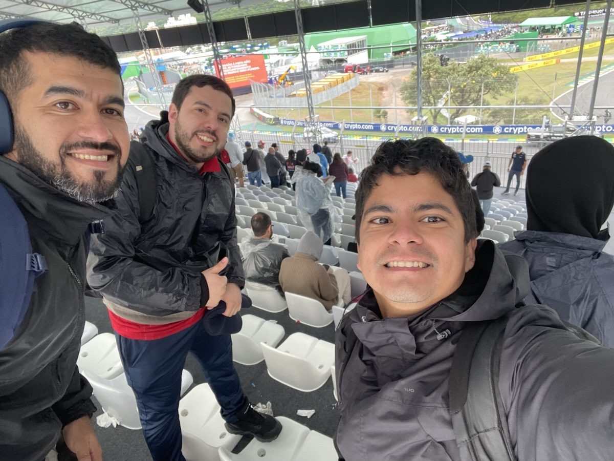 Tres viajeros de JUHU Travel sonriendo en una tribuna de Interlagos, con la pista detrás