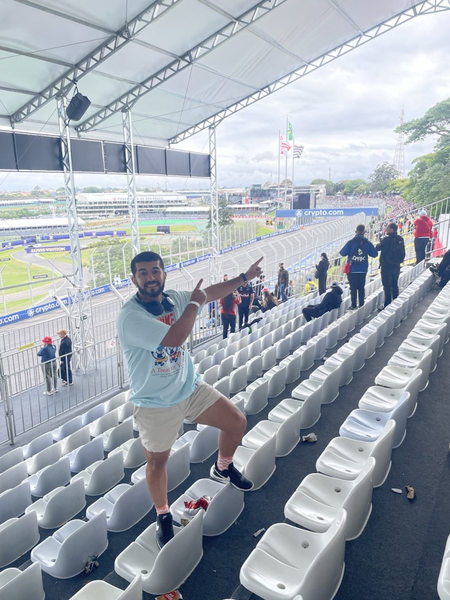 Un viajero de JUHU Travel de pie entre las butacas de una tribuna techada de Interlagos, con el circuito y São Paulo abriéndose al fondo