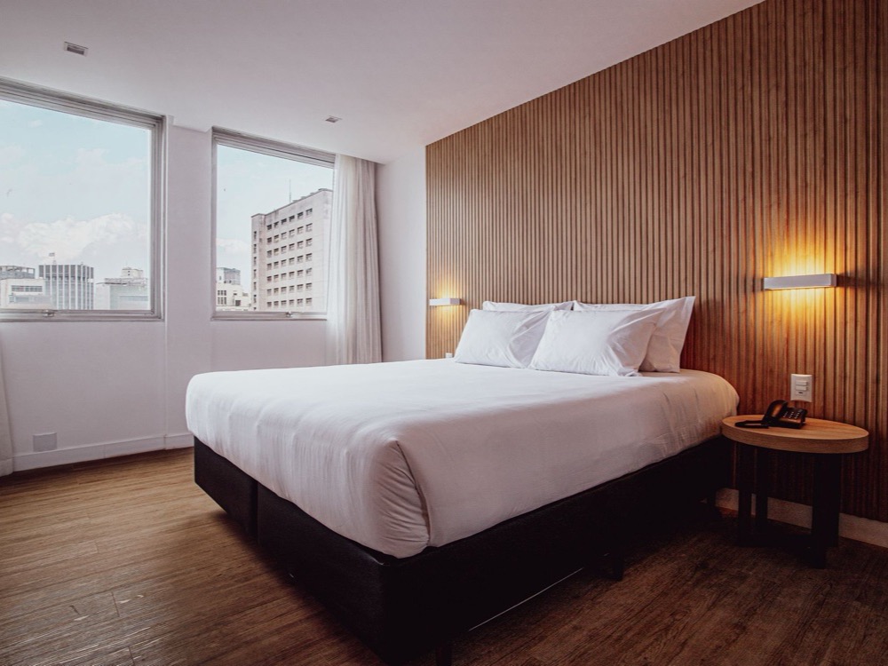 Habitación del Sooz Hotel Collection con cama matrimonial de ropa blanca, pared de listones de madera con dos luces, mesa de luz y ventanas
