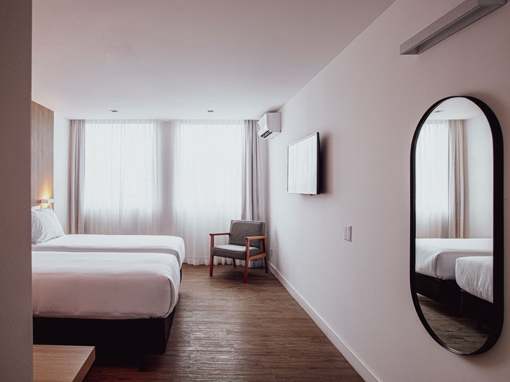 Habitación del Sooz Hotel Collection con dos camas individuales de ropa blanca, un sillón junto a la ventana, TV en la pared y un espejo ovalado